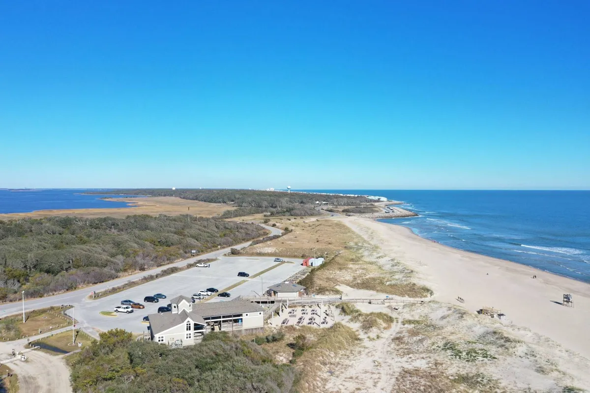 The image shows an expansive beach with calm ocean waves, a distant lighthouse, and accessible nature trails stretching along the coast. The inviting sandy shoreline suggests a variety of group-friendly activities from sunbathing to outdoor dining.