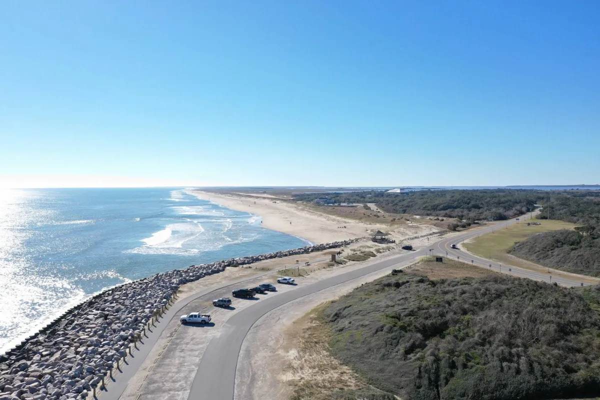 A tranquil view of the nearby beach with the ocean, jetty, and parked cars visible. The shoreline offers a sense of proximity to the beach, reinforcing the narrative of returning home after island adventures.
