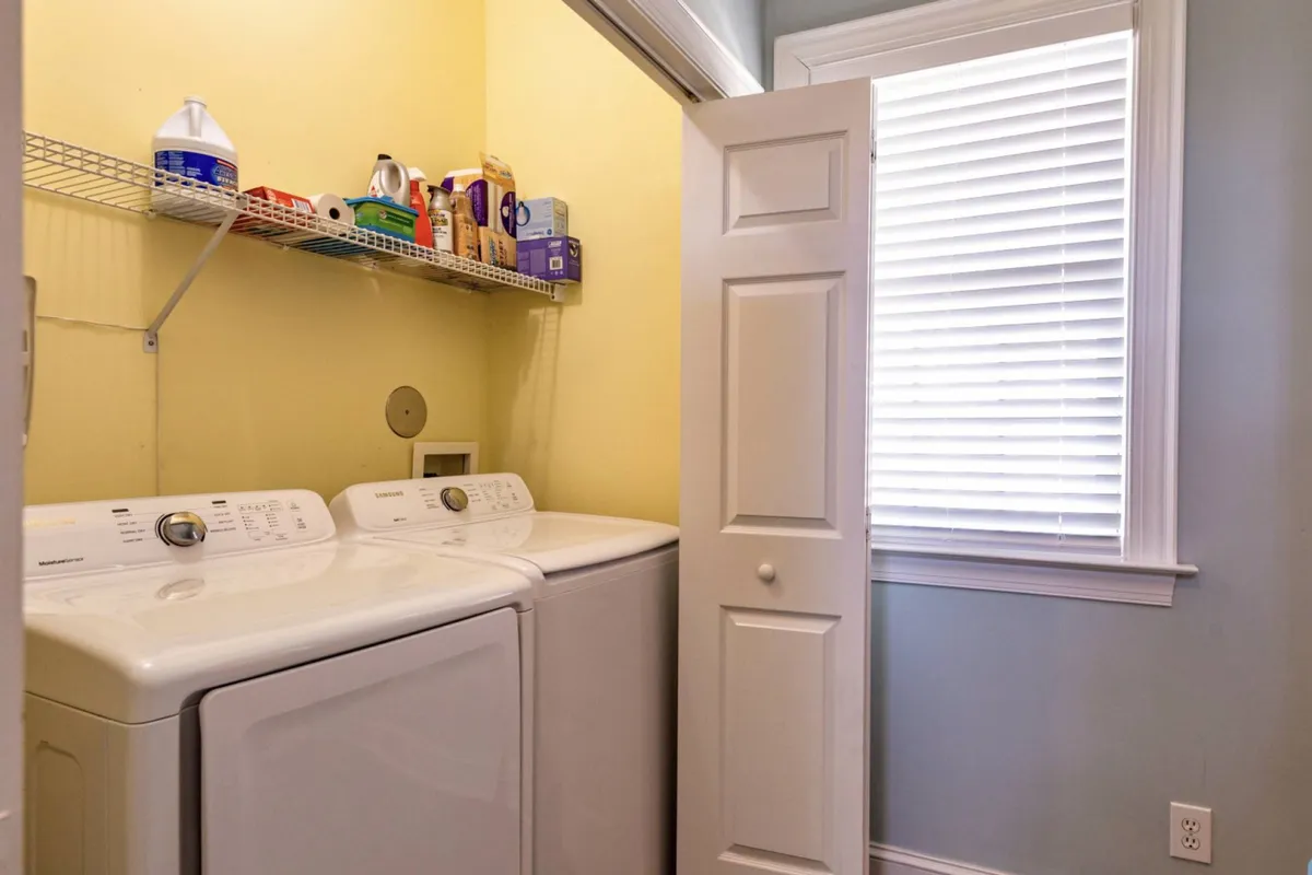 A functional laundry area with a washing machine, dryer, and storage shelves stocked with laundry supplies. The window allows in natural light, highlighting the room's practical features.