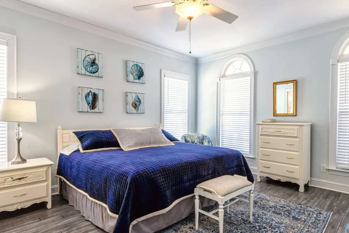 A serene bedroom featuring a comfortable bed with crisp linens, decorative art on the walls, a lamp on the nightstand, and a dresser. Natural morning light filters through the window, creating a rejuvenating atmosphere perfect for relaxing or reading.