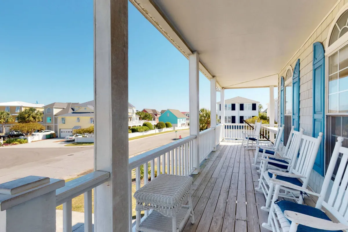 A classic wooden wraparound porch with inviting rocking chairs, blue shutters, and a view toward the street. The space feels both cozy and connected to the outdoors, perfect for relaxing or socializing as ocean breezes flow in.