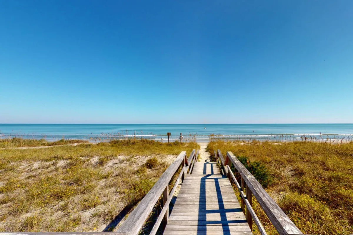 A scenic wooden walkway stretches through grassy dunes, leading directly to a wide, sandy beach with the ocean glimmering in the distance. The scene exudes tranquility and highlights the ease of direct beach access. Soft morning light and gentle waves evoke an inviting, serene coastal atmosphere.