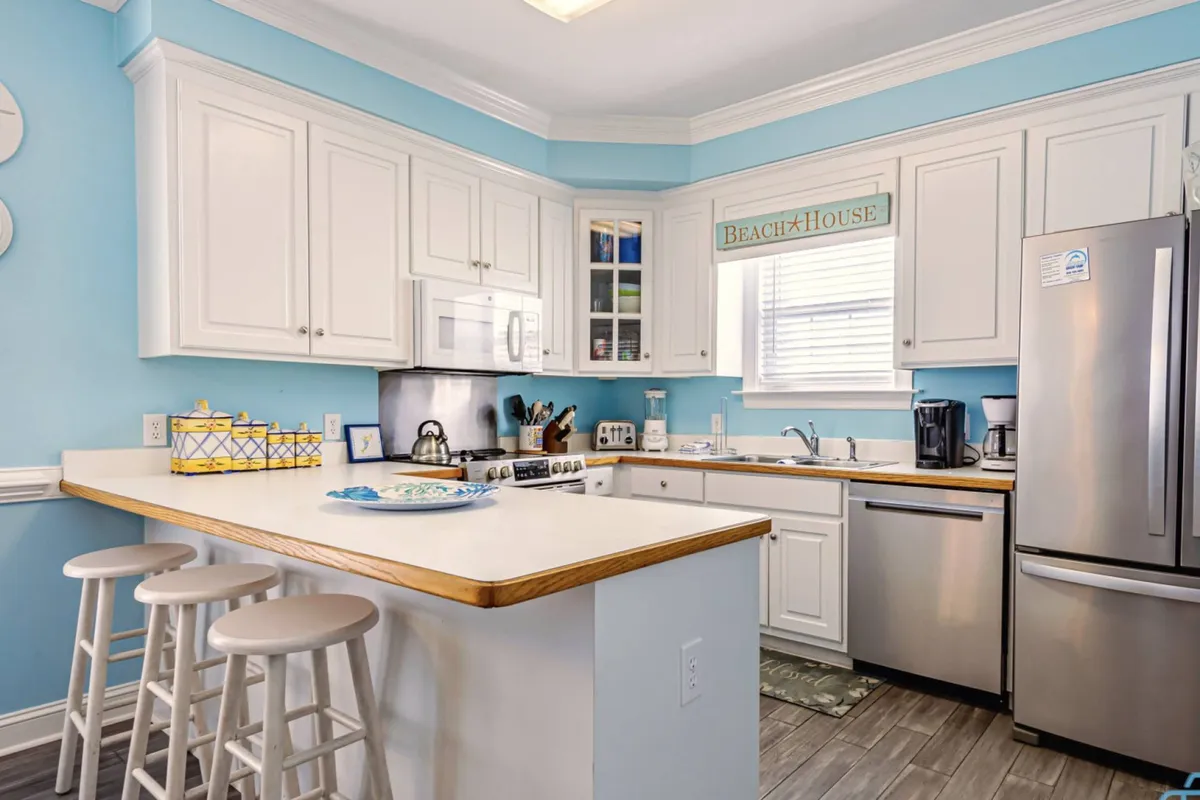 A bright, welcoming kitchen highlighted by light blue walls, white cabinetry, and a kitchen island with bar stools. Beach-themed decor adds a coastal touch, while the setup is clearly equipped for cooking and entertaining, encouraging groups to gather and enjoy meals together.