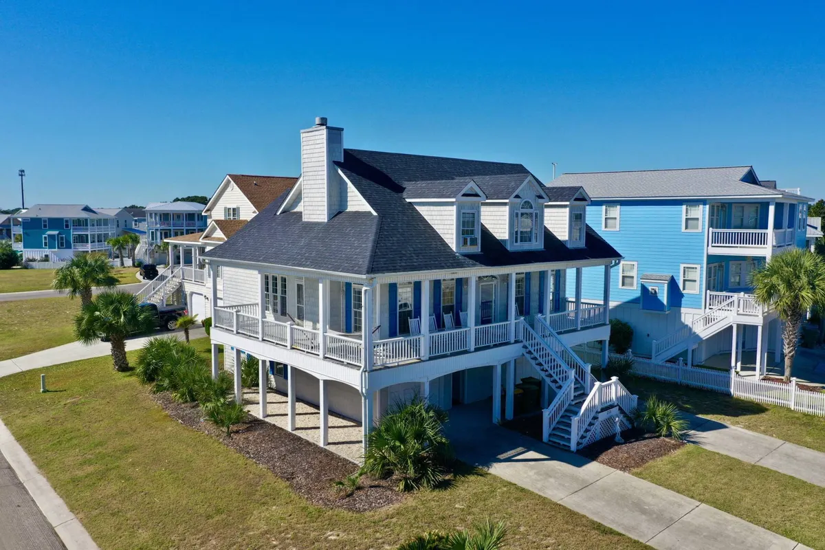 The image showcases the charming exterior of The Hamptonian, a three-level single-family home in Kure Beach. The focal points include a spacious wraparound front porch perfect for relaxing, classic dormer windows that add coastal character, and a beautifully landscaped yard. The inviting porch is set up for enjoying sunset views, ocean breezes, and family gatherings.