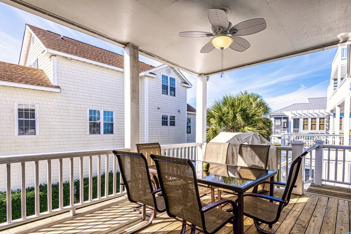 A welcoming outdoor dining area with a table set on a shaded porch, complete with a ceiling fan, surrounded by palm trees and neighboring homes. The space is designed for comfort and group gatherings, suggesting a perfect setting for outdoor meals and casual relaxation.