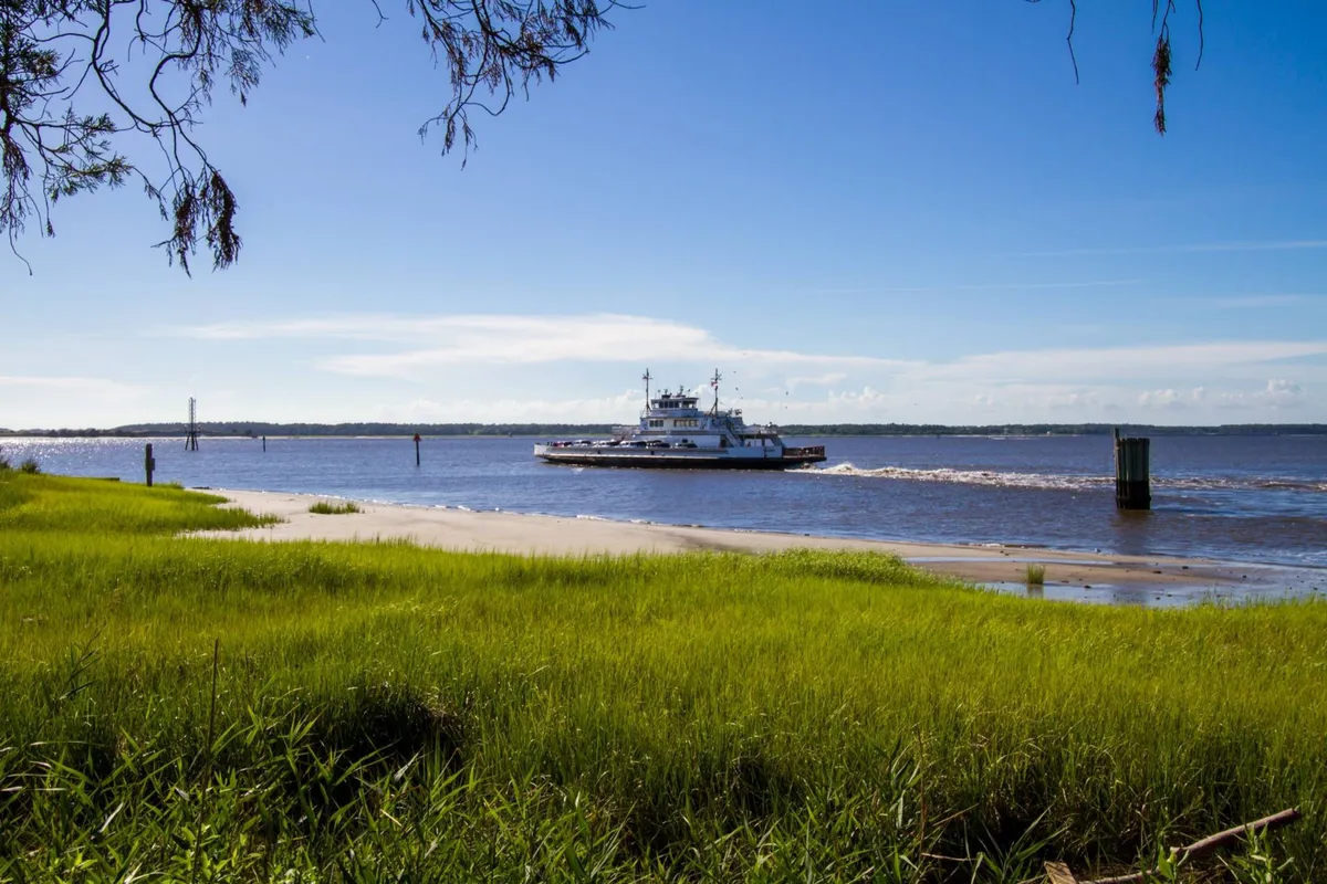 A scenic view of a ferry boat gliding along a river bordered by lush green grass under a blue sky. The tranquil setting suggests boating, fishing, and nature walks, with a sense of adventure and the outdoors.