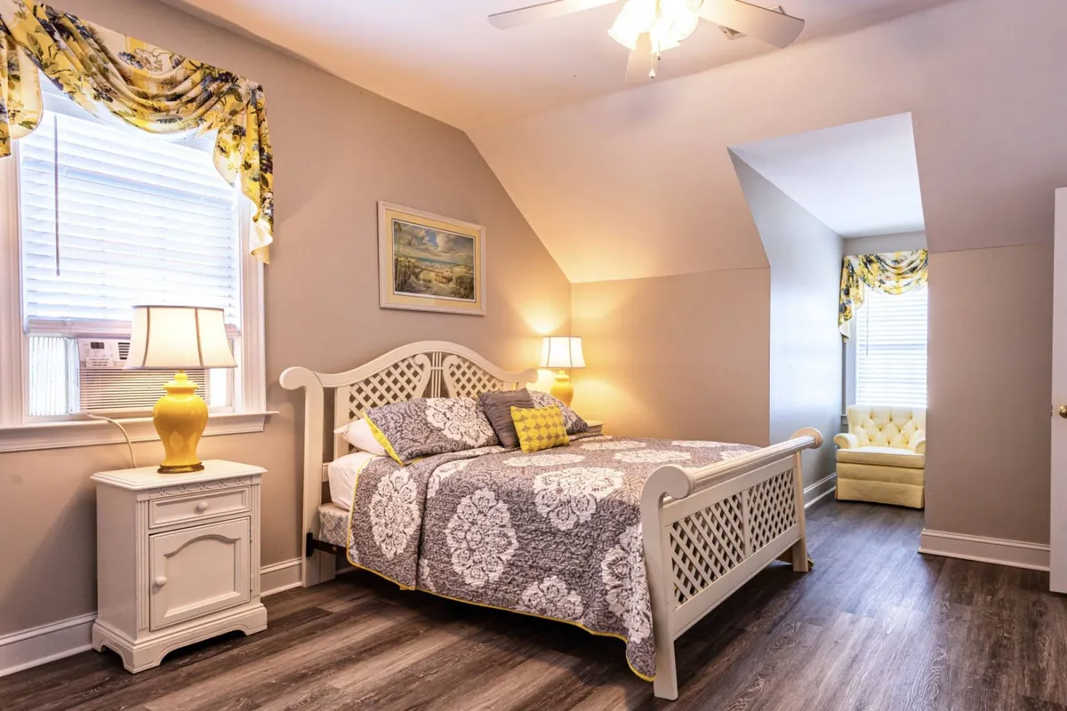 A spacious bedroom with a plush bed, an armchair by the window, a nightstand, and soft window treatments. The setting invites relaxation, with artwork adding a touch of style and comfort.