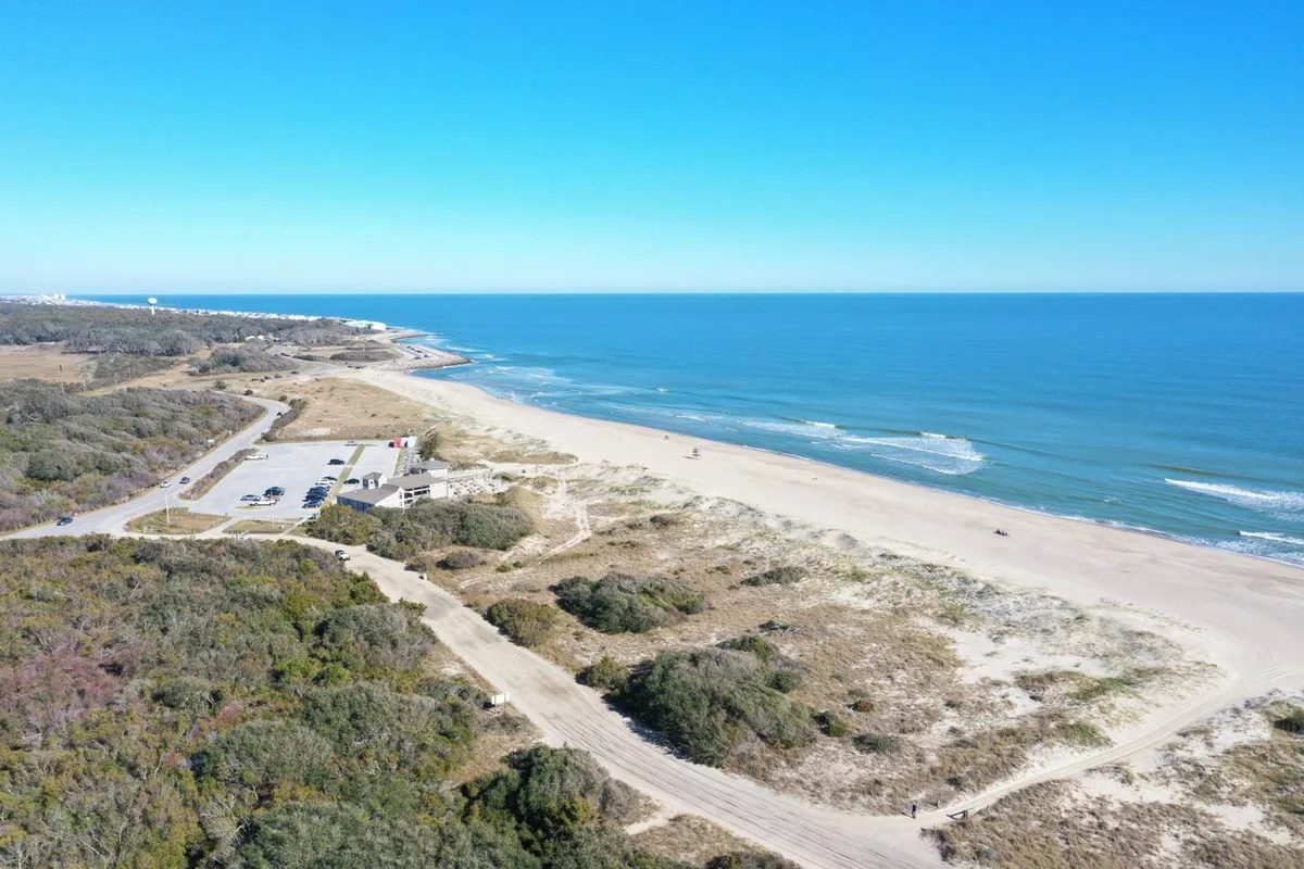 A relaxed exterior shot of the beach and ocean with a parking area and grassy dunes in the foreground. The laid-back beach vibe is accented by open space and hints of a welcoming community.