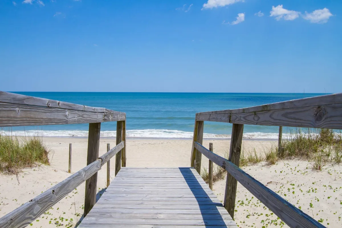 A wooden walkway opens directly onto a pristine, sandy beach with gentle ocean waves under a wide, blue sky. The path is framed by beach grasses, reinforcing the connection from home to shore and the secluded, natural feel of the area.