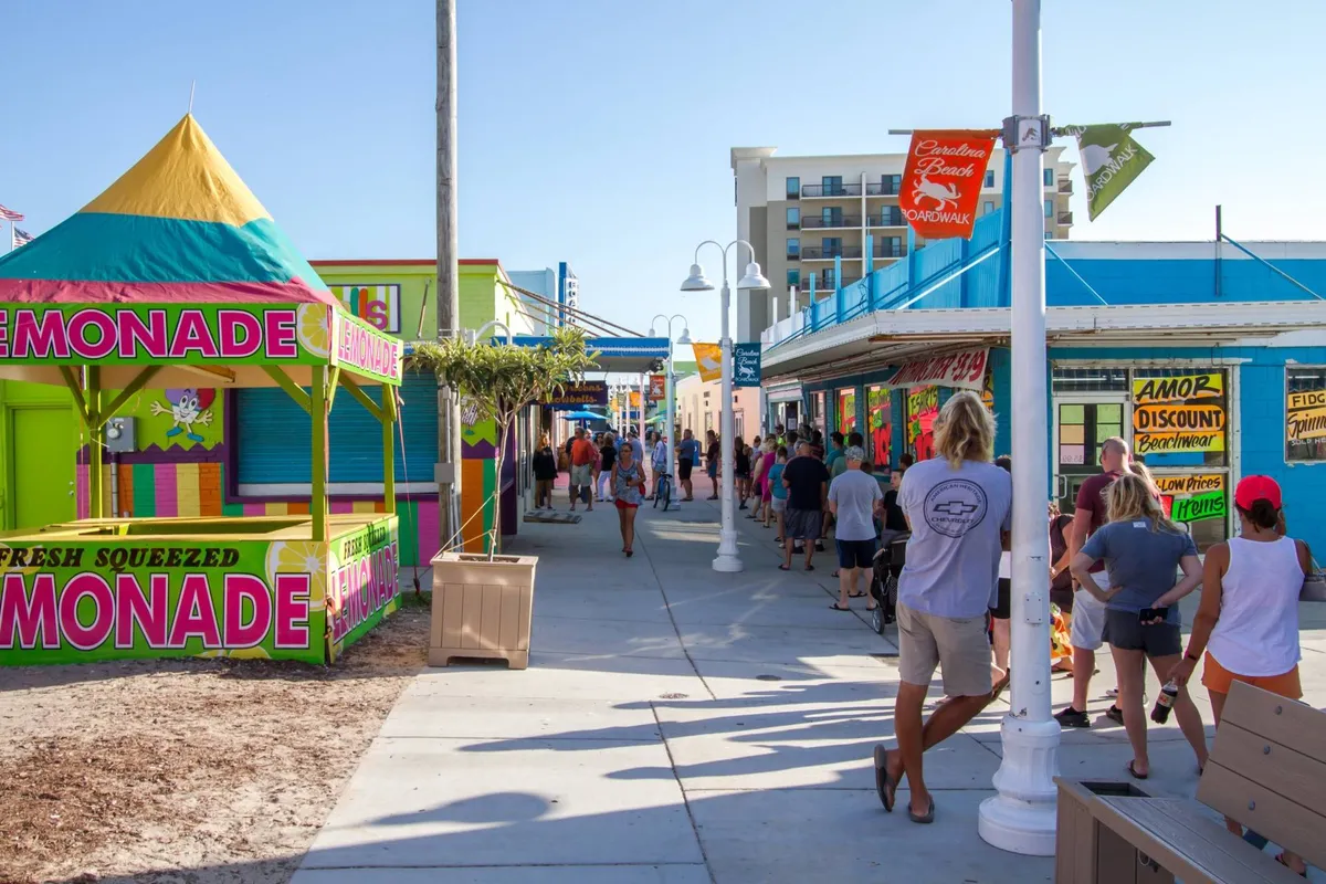 This vibrant exterior image features a colorful lemonade stand and brightly colored storefronts, with people walking and enjoying the lively atmosphere. Decorative signage and the festive mood evoke the energetic spirit of a boardwalk area.