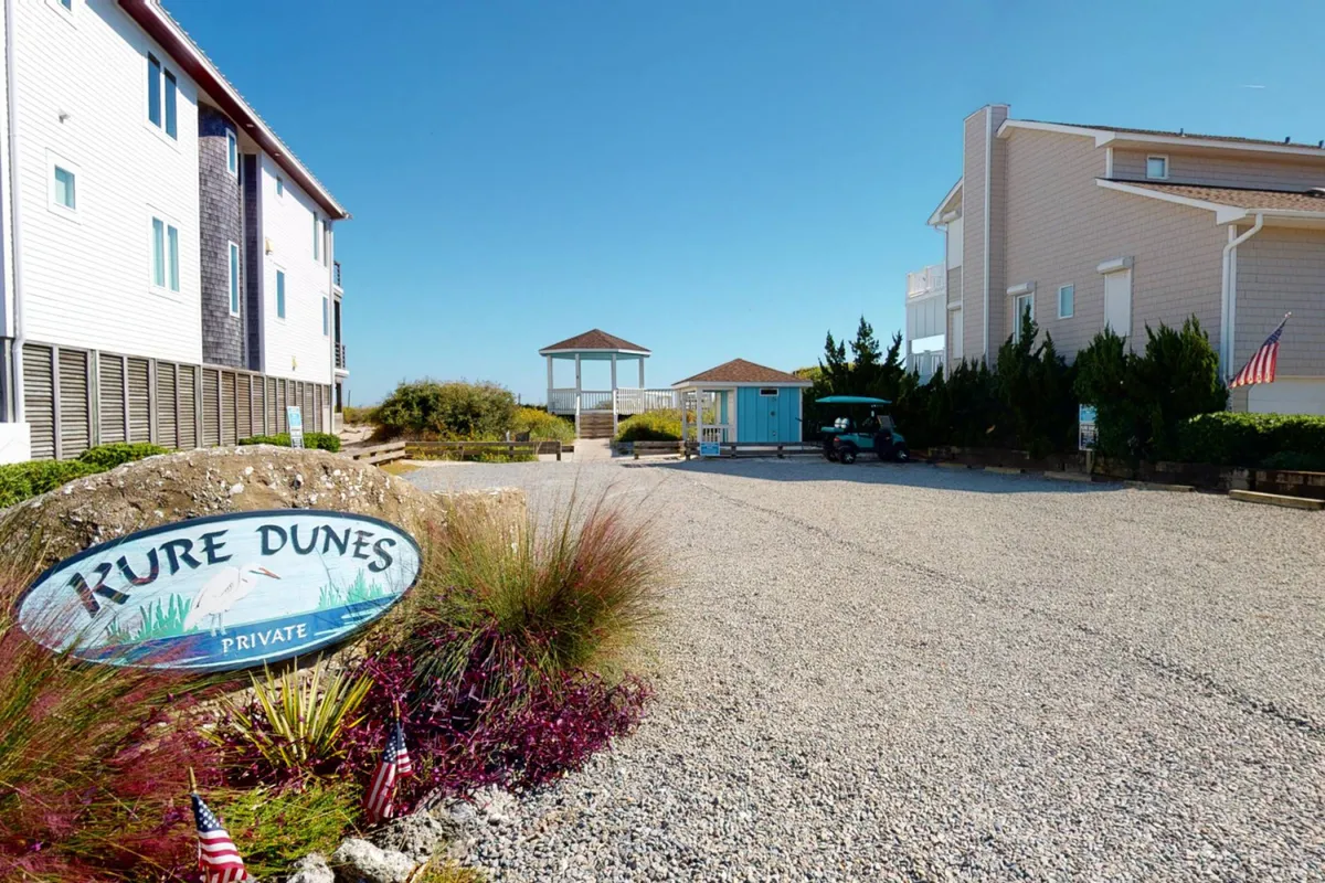 The exterior view of the Kure Dunes neighborhood, showing a sign, gravel driveway, gazebo, blue building, and lush greenery. The area invites guests to explore the outdoors with accessible walking paths and green spaces.